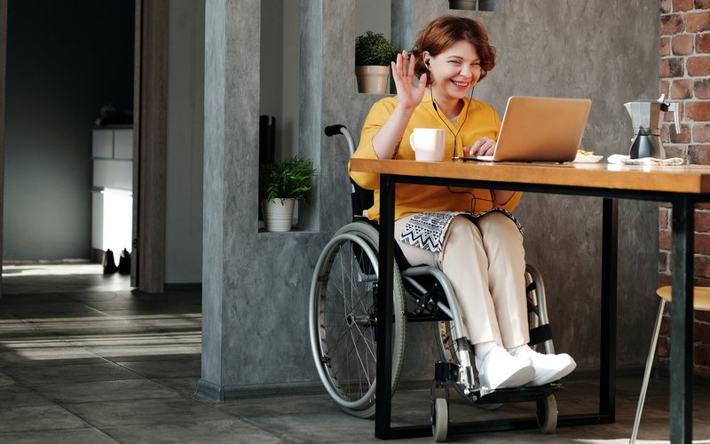 Photo of a woman sitting in a wheelchair smiling waving and using a laptop