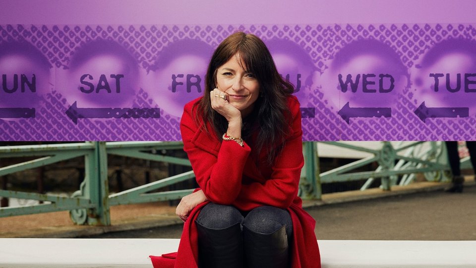 Davina McCall in a red coat sitting and facing camera with head in hand. Behind her is a big billboard of a purple pill packet.