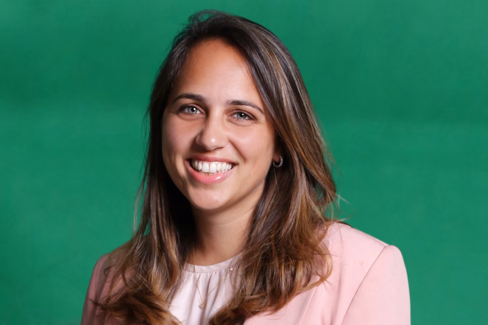 Dr Melanie Nana is wearing a light pink blazer and shirt and smiling broadly at the camera against a dark green backdrop