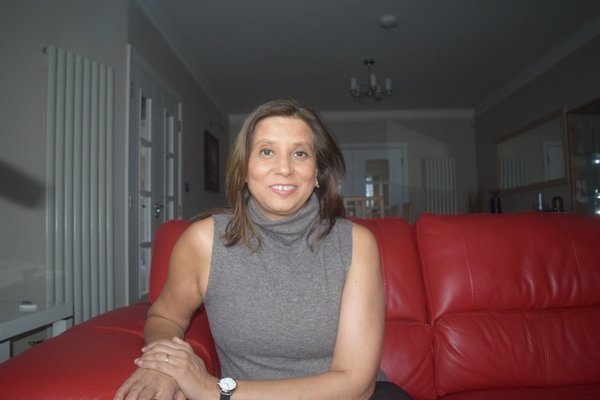 An Asian women wearing a grey sleeveless turtle neck jumper, sitting on a red sofa - smiling to the camera.