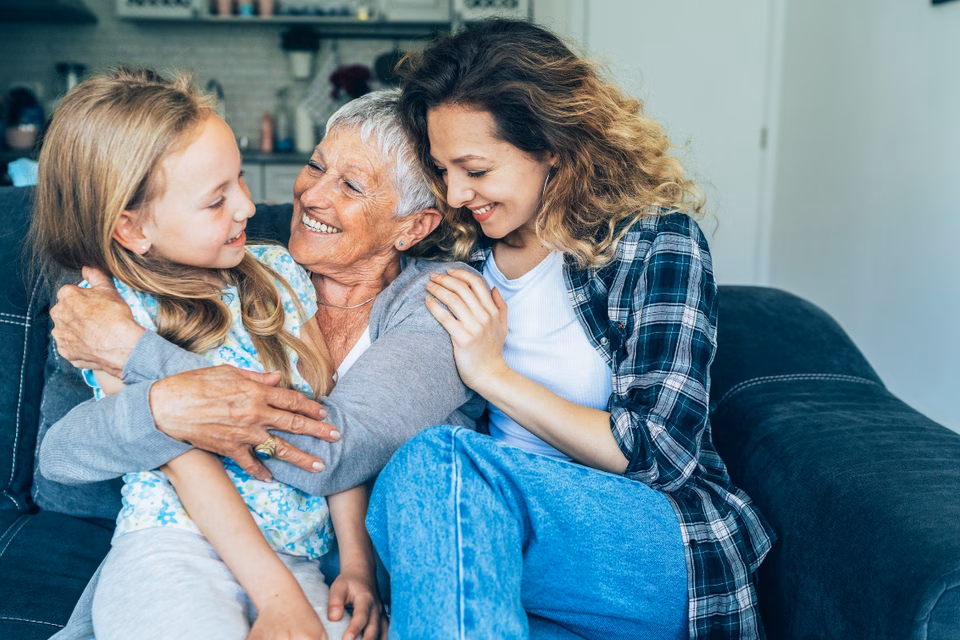 A blonde girl being hugged by her grandma and mum all smiling sat on a couch