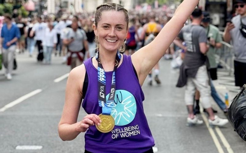 Researcher Marianne Watters holds a running medal smiling in Wellbeing of Women t-shirt
