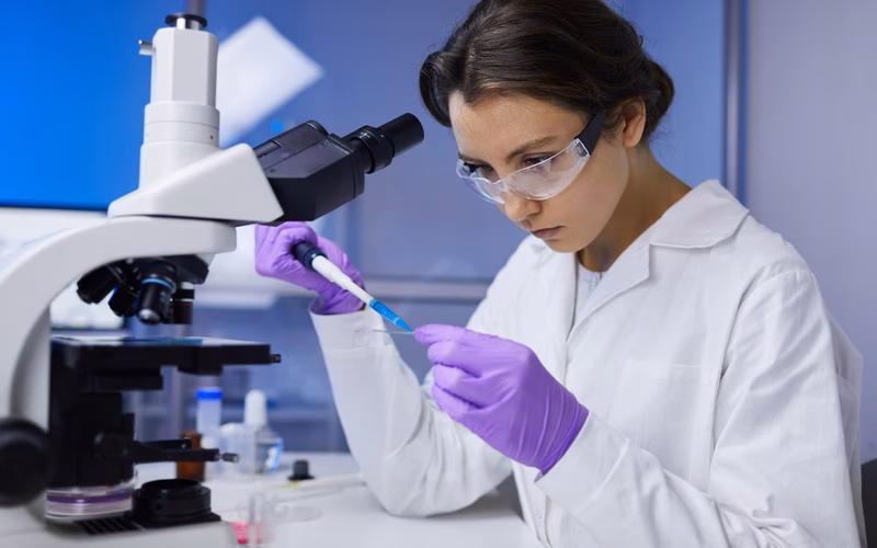 A young female researcher adds substance to a slide to study under a nearby microscope