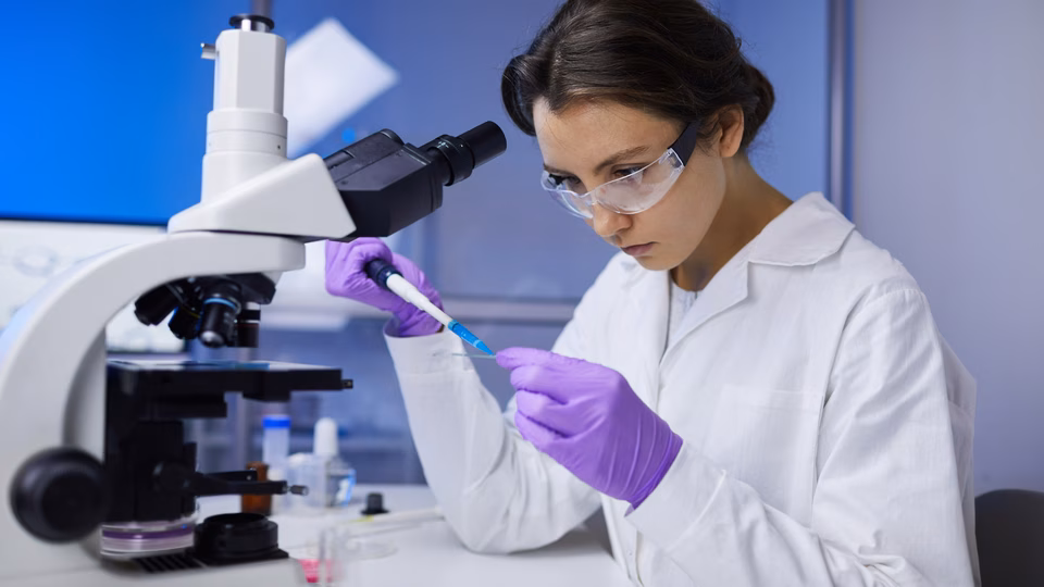 A young female researcher adds substance to a slide to study under a nearby microscope