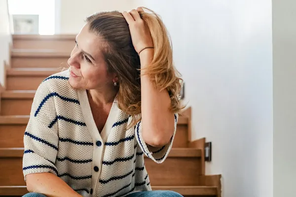 A woman sitting on a wooden staircase, wearing a cream-and-navy striped cardigan, looking to the side while holding her ponytail.